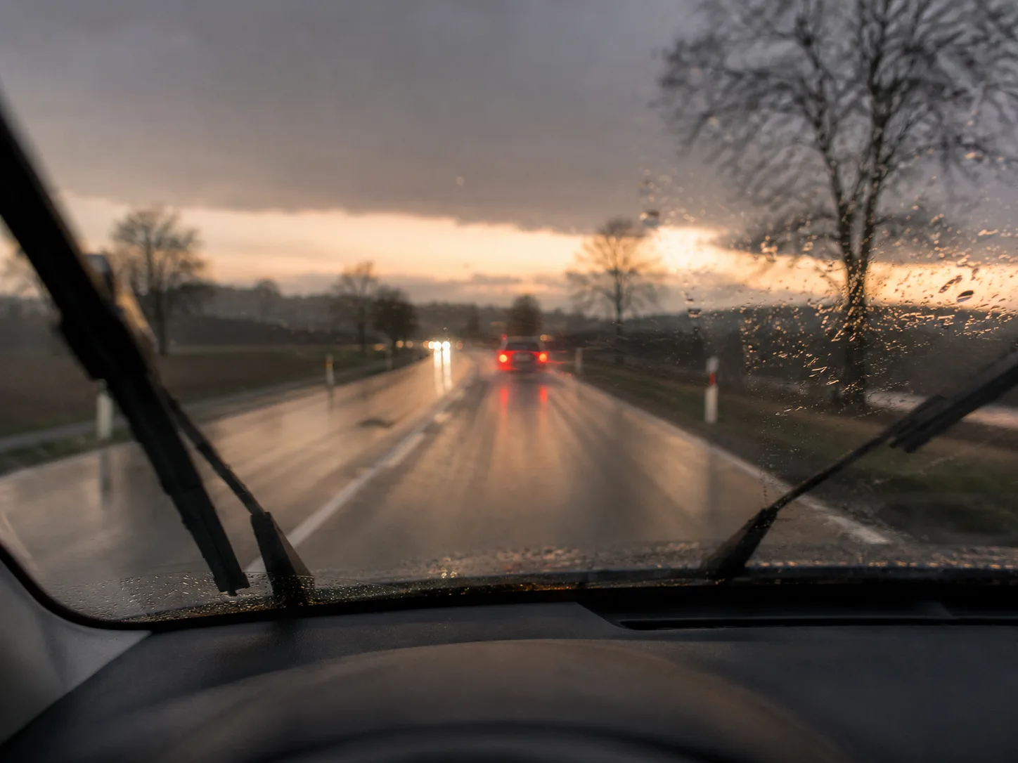 Blick durch die Windschutzscheibe auf eine nasse Landstraße bei Dämmerung, Rücklichter voraus